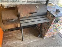 Full front view of vintage charcoal barbecue grill showing main cooking area, smoker box, wooden slat front shelf, and colorful sticker-covered side cabinet with wheels.