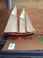 Front-view of vintage wooden model ship on a wooden stand, showing two masts with white fabric sails and detailed rigging.