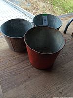 Three vintage sap buckets viewed from above placed on a wooden surface, showing metal interior and red painted exterior on one bucket.