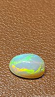 Oval cabochon cut natural Ethiopian opal showing bright green and yellow full rainbow flash colors on a brown textured background.