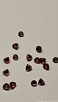 Multiple natural Almandine garnet gemstones scattered on a neutral background, showing their small size, bullet cut, and rich reddish-brown color.