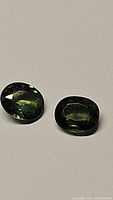 Pair of oval cut green sapphires showing faceting and dark green color on flat surface