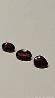 Three deep red garnet gemstones of mixed cuts (oval and pear) shown reflecting light and displaying facets.
