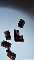 Six deep red rectangular baguette cut almandine garnet gemstones on white background showing rich color and clarity.