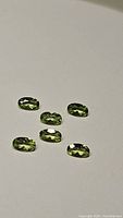 Six natural apple green peridot oval cut gemstones approximately 5x3mm sized laid on white surface.