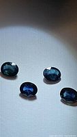 Four oval cut natural blue sapphires with visible inclusions laid out individually on a white surface, showing color and cut under light.