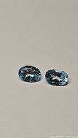 Two oval shaped dark blue natural topaz gemstones lying side by side on a plain background, showing faceted cuts and excellent clarity.