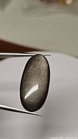 Close-up photo of the oval cabochon Silver Sheen Obsidian gemstone held by tweezers showing its shape, color, and silver flash effect under light.