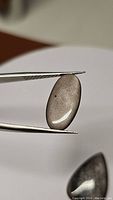 Close-up photo of one oval cabochon Silver Sheen Obsidian gemstone held by tweezers, showing smooth polished surface and reflective silver sheen.