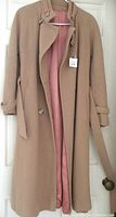 Full view of the tan ladies winter coat hanging showing belted waist, button, and pink lining.