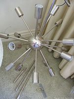 Photo of light fixture showing central chrome sphere with 12 metal arms extending outward with cylindrical bulb holders, lay on carpet with some parts disassembled.