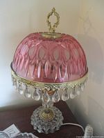 Full view of pink glass dome shade with clear crystal prisms and ornate brass finial and band, standing on clear glass base.