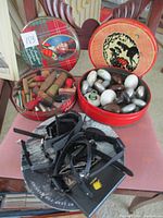Two vintage round cookie tins filled with various items on a table. Left tin has wooden stamps, right tin has metal and ceramic door knobs and a black smart watch on top.