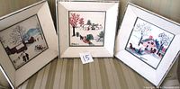 Three folk art pictures signed Moses showing winter scenes with houses, trees, sledding, and people outdoors, framed and placed on a striped background.