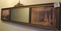 Full view of the long rectangular framed piece showing a mirror in center flanked by Egyptian scene art prints inside a carved wooden frame.