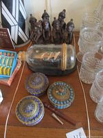 Lot of Native tribal figural cast metal napkin rings, Tibetan covered tins, and ship in a bottle on table.