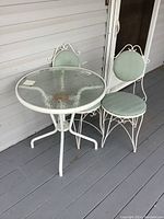 Full view of metal framed glass top table and two matching chairs with green seats on porch floor.