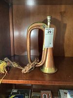 Copper and brass bugle with a decorative tassel cord placed on a wooden shelf, showing side view and condition with slight dents and patina.
