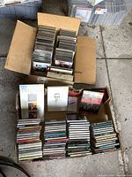Two cardboard boxes filled with CDs and some cassette tapes arranged vertically and horizontally inside. Visible album covers include The Who, John Cougar Mellencamp, and others.