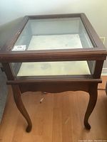 Full front view of the wooden display table with glass top, showing curved legs and visible scratches on the wood surface.