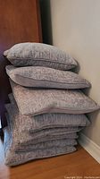 Stack of eight grey plush accent pillows leaning against a wall and wooden furniture showing their texture and shape.