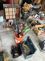 Overview photo of fishing gear bucket, showing multiple fishing poles with reels standing inside an orange bucket along with two tackle boxes and other miscellaneous items on garage floor.