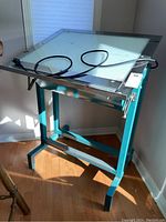 Photo showing the light table from a front angle, highlighting the metal frame, adjustable top, and power cord coiled on the surface.