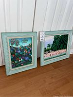 Two framed Caribbean style paintings set on the floor against a wall, showing overall presentation and frame styles.