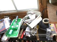 Wide shot showing stacked and grouped painting supplies including trays, brushes, sandpaper, and tape on a cluttered surface near a window.