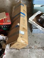 Full box containing folded party tent, secured with tape, placed on concrete floor next to other boxes.