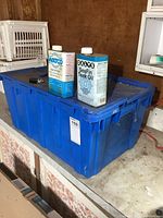Two nearly full cans of teak oil, Watco and Dalys SeaFin, placed on top of a blue plastic tote box.