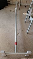 Full view of the white adjustable metal shower safety pole extended, showing the main pole and crossbar base with rubber feet covers on carpeted floor.