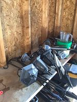 Photo showing a collection of black metal hexagonal solar lantern garden lights with clear panels and spike stakes along with long driveway marker lights with translucent round tops, placed leaning against a wooden surface.