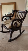 Side view of dark mahogany finish rocking chair with beige cushion on floor. Shows curved armrest detail and leg carving.