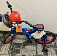 Full view of the children's bike featuring Spider-Man themed graphics, a red mask on front basket, training wheels, and seat with spider emblem.