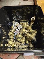 Close-up photo of black container filled with various new brass fittings including valves, connectors, T-shaped, and elbow fittings.