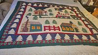 Full view of the quilt showing the Christmas village design with colorful houses, trees, and a train, bordered with red and green fabric and white tree shapes.