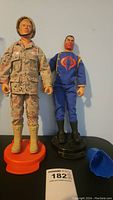 Front view of two GI Joe figures with one in desert camouflage and the other in blue outfit along with two display stands and a blue accessory piece.