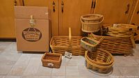 Photo of all baskets showing 4 Peterboro baskets with tags and 3 other baskets on the floor in front of wooden cabinetry.