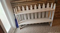 Full frontal view of white wooden picket fence shelf unit showing two shelves and vertical pointed picket slats.