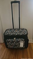 Full frontal view of portable sewing bag with patterned top and front zipper compartments, wheels and handle visible.