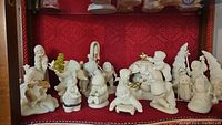 Front angle of approx. 13 white porcelain Snow Babies figurines arranged on shelf with red background.