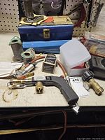 Soldering supplies including wire spools, torch soldering gun with nozzle, boxes of solder, blue toolbox, and small container arranged on workbench.