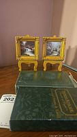 Two gold miniature frames on easels displayed on a table with original boxes in foreground