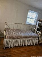 White metal trundle day bed with gold spherical finials at the four corners and floral design on frame, mattress on top, lace skirt around trundle area, wooden floor beneath.