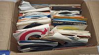 Over 200 45 RPM records stacked in a cardboard box with visible worn paper sleeves in mixed condition.