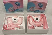 Two boxed kids digital cameras with pink cat designs, displayed with packaging in the background showing the product box.