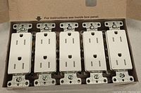 Photo of five white plug outlets in a cardboard box showing the front faces with two vertical slots and grounding hole.