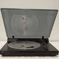 Front angled view of AKAI AP-A150 turntable with dust cover open. Shows metal platter with cutouts, tonearm, base, and AKAI branding.
