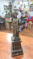 Front angle of the brass Statue of Liberty figurine placed on a wooden surface showing detailed construction of torch, crown, and pedestal base.